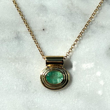 A gold chain necklace with a round bezel-set green gemstone pendant, displayed on a white marble surface.